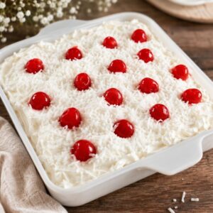 Creamy no bake ice box cake in a white baking dish topped with shredded coconut and maraschino cherries. The easy layered dessert is displayed on a rustic wooden table with a soft neutral cloth and decorative flowers in the background.